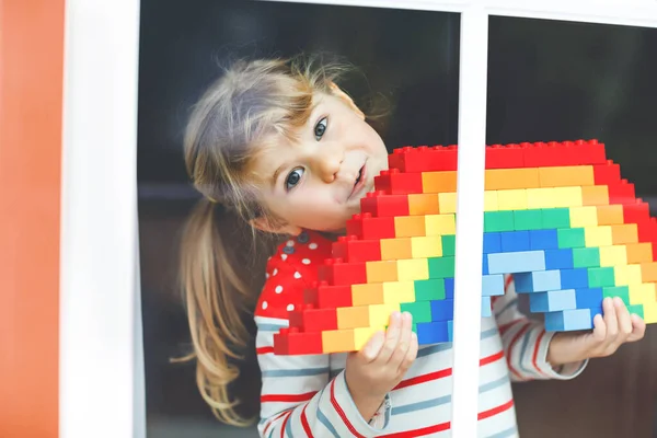 Cute little toddler girl by window create rainbow with colorful plastic blocks during pandemic coronavirus quarantine. Children made and paint rainbows around the world as sign. — Stock Photo, Image