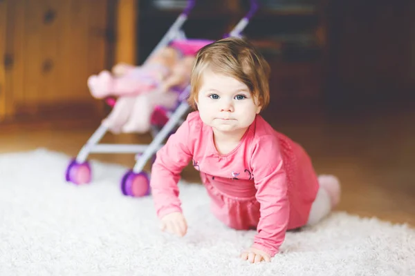 Schattig schattig baby meisje kruipen en spelen met pop rijtuig. Prachtig peuterkind met kinderwagen met kinderspeeltje thuis. Gelukkig kind met twee poppen — Stockfoto