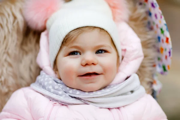 Nettes kleines, hübsches Mädchen, das an kalten Herbst-, Winter- oder Frühlingstagen im Kinderwagen oder Kinderwagen sitzt. Glücklich lächelndes Kind in warmer Kleidung, modisch stylischem Babymantel und Hut. Schnee fällt herunter — Stockfoto