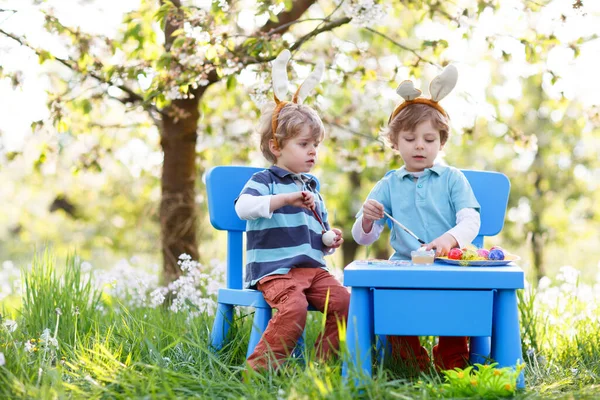 Two cute little kid boys wearing Easter bunny ears, painting colorful eggs and having fun outdoors. Family, siblings, brothers, twins celebrating religious holiday. Best friends coloring eggs. — Stock Photo, Image
