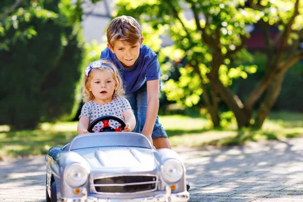 Dwoje szczęśliwych dzieci bawiących się dużym starym samochodzikiem w letnim ogrodzie, na świeżym powietrzu. Chłopak prowadzi samochód z małą dziewczynką, urocza siostra w środku. Śmiejące się i uśmiechnięte dzieci. Rodzina, dzieciństwo, koncepcja stylu życia. — Zdjęcie stockowe