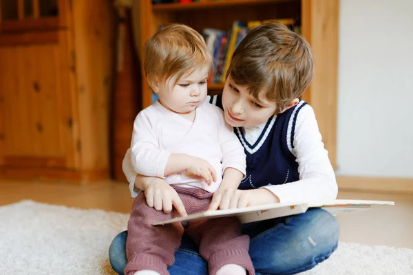 En skolegutt som leser bok for småbarnsjente, to søsken som sitter sammen og leser bøker. Vakker familie i kjærlighet, søt baby og barn som har det gøy hjemme, inne. – stockfoto