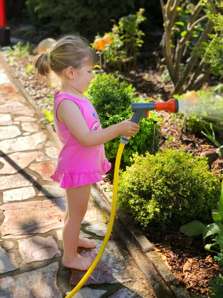 Belle petite fille tout-petit arrosant des fleurs de jardin avec tuyau d'eau le jour d'été. Enfant heureux aidant dans le jardin familial, à l'extérieur, s'amusant avec éclaboussures — Photo