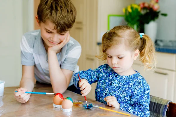 Opgewonden kleine peuter meisje en oudere broer, schooljongen kleuren eieren voor Pasen. Twee schattige kinderen, broers en zussen op zoek verrast naar kleurrijke eieren, vieren vakantie met familie. — Stockfoto