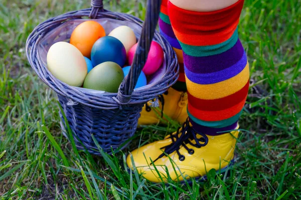 Close-up van de benen van peuter meisje met kleurrijke kousen en schoenen en mand met gekleurde eieren. Kind heeft plezier met traditionele paaseieren jacht, buiten. Viering van christelijke feestdagen. — Stockfoto