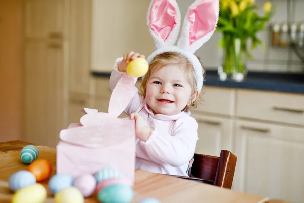 Roztomilé malé batole dívka na sobě velikonoční zajíček uši hrát s barevnými pastelovými vejci. Šťastné dítě vybaluje dárky. Rozkošné zdravé usměvavé dítě v růžových šatech těší rodinnou dovolenou — Stock fotografie