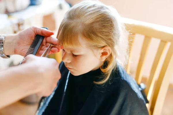 Adorable toddler girl getting his first haircut. Happy cute child sitting at the barbershop. Hairdresser making a hair style to cute little baby girl — Stock Photo, Image