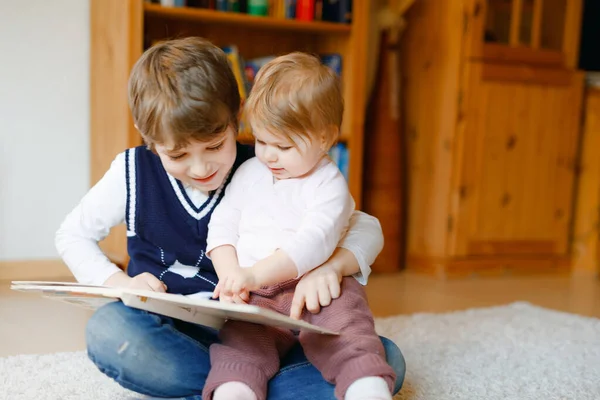 School kid boy reading book for little toddler baby girl, Two siblings sitting together and read books. Beautiful lovely family in love, cute baby and child having fun at home, indoors. — Stock Photo, Image
