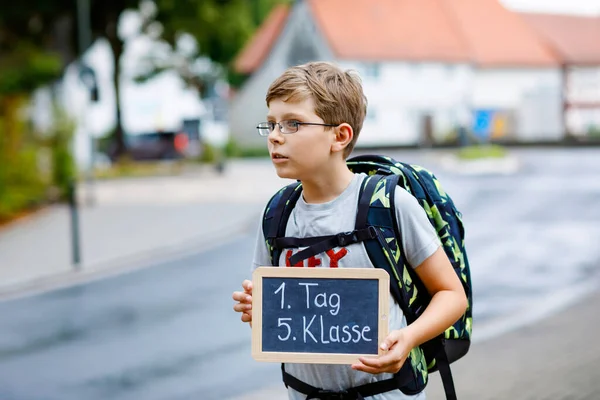 Ragazzino felice con occhiali e zaino o borsa. Studente sulla strada per la scuola media o superiore. Bambino all'aperto per strada. Torniamo a scuola. Sulla scrivania Primo giorno quinta elementare in tedesco. — Foto Stock