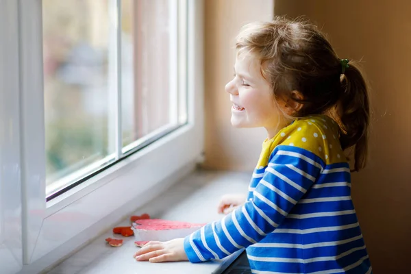 Nettes kleines Mädchen am Fenster mit vielen Teigherzen als Geschenk zum Valentinstag, Muttertag oder Geburtstag. Entzückend glücklich lächelndes Kind drinnen. — Stockfoto