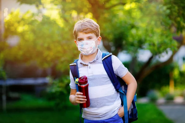 Rapazinho feliz, máscara médica, garrafa de água e mochila ou mochila. Um estudante a caminho da escola. Criança saudável ao ar livre. De volta à escola após o tempo de quarentena do bloqueio da doença pandêmica da corona — Fotografia de Stock