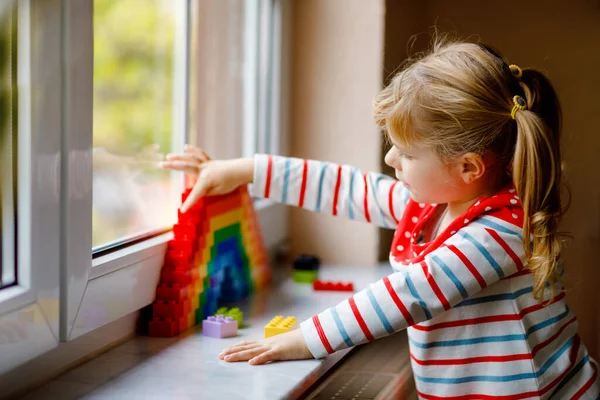 Söt liten småbarn flicka genom fönstret skapa regnbåge med färgglada plastblock under pandemisk coronavirus karantän. Barn gjorde och måla regnbågar runt om i världen som tecken. — Stockfoto