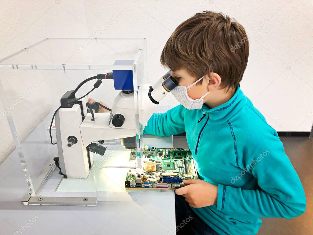 Kid boy conducts experiment with microscope in school lab. Curious inquisitive child learning physics and computer scheme. Schoolboy with medical mask due to coronavirus pandemic.