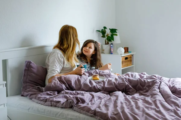 Belle jeune mère et mignonne écolière câlinant ensemble au lit le matin. Famille heureuse de jeune femme célibataire et belle fille prendre le petit déjeuner, manger sur la fête des mères. — Photo