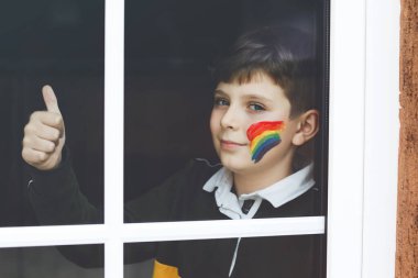 Yalnız okul çocuğu pencerenin kenarında oturuyor yüzü renkli gökkuşağıyla. Çocuk pandemik koronavirüs karantinasında. Çocuklar dünyanın her yerinde gökkuşağı yapar ve boyarlar.