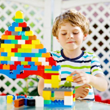 Renkli plastik bloklarla oynayan küçük sarışın çocuk. Sevimli kreş çocuğu renkli tişörtler giyiyor ve büyük bir kale inşa edip bir ev yaratarak eğleniyor.