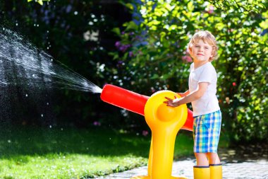 Sıcak ve güneşli bir yaz gününde bahçe hortumuyla oynayan komik çocuk. Çocuk su serpme ve yağmur damlalarıyla eğleniyor. Çocuklar için açık havada su ile vakit geçirmek. Lastik çizmeler..