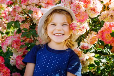 Çiçek açan gül bahçesinde küçük bir kızın portresi. Yaz güneşli bir günde parkta güllerle ve çiçeklerle eğlenen sevimli güzel bir çocuk. Mutlu gülümseyen bebek.