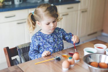 Heyecanlı küçük kız Paskalya için yumurta boyuyor. Sevimli mutlu çocuk renkli yumurtalara şaşırmış görünüyor, tatilini ailesiyle kutluyor. Evde farklı renkte sevimli bir çocuk var..