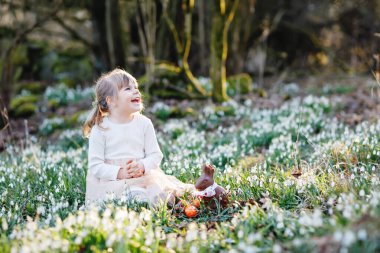 Güneşli bir günde, açık havada bahar ormanlarında Paskalya yumurtası avına çıkan küçük kız. Bir sürü kartopu çiçeği ve çikolatalı tavşanı olan sevimli mutlu bir çocuk. İlkbahar, Hıristiyan bayramı konsepti.