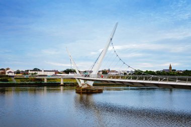 Derry, İrlanda. Kuzey İrlanda 'da, Derry Londonderry' de, Kültür Şehri 'nde şehir merkezinde aydınlatılmış bir barış köprüsü. Gece bulutlu gökyüzü, alacakaranlıkta nehirde yansıyor.