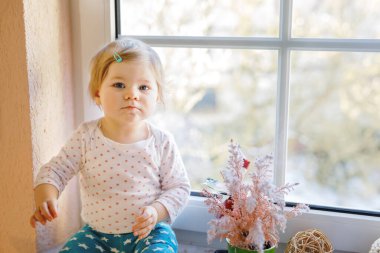 Mutlu sevimli kız bebek pencerenin yanında oturuyor ve kışın veya ilkbaharda dışarıda karları seyrediyor.