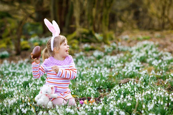 Little girl with Easter bunny ears making egg hunt in spring forest on sunny day, outdoors. Cute happy child with lots of snowdrop flowers, huge chocolate egg and colored eggs.