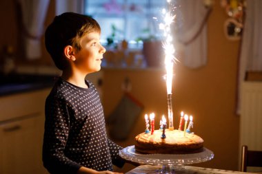 Sevimli, mutlu, küçük sarışın çocuk doğum gününü kutluyor. Çocuk ev yapımı kek üzerine mum üflüyor, kapalı alanda. Okul çocukları için doğum günü partisi, aile kutlaması, havai fişekler.