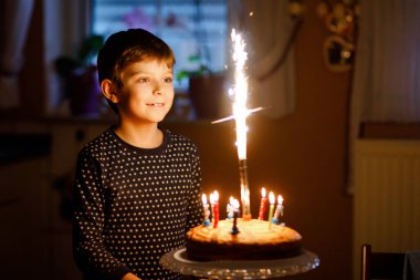 Sevimli, mutlu, küçük sarışın çocuk doğum gününü kutluyor. Çocuk ev yapımı kek üzerine mum üflüyor, kapalı alanda. Okul çocukları için doğum günü partisi, aile kutlaması, havai fişekler.