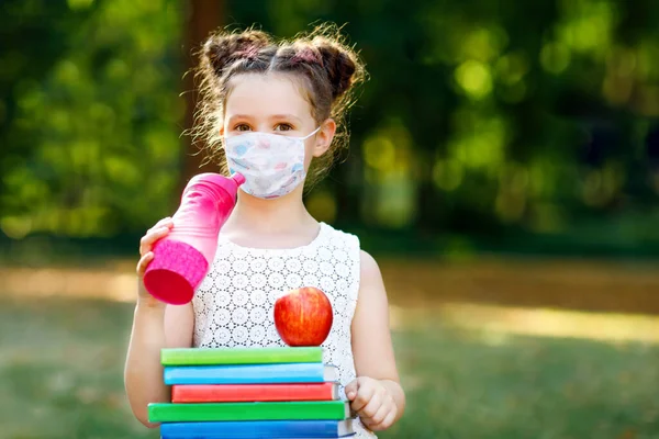 Happy little kid girl with medical mask, water bottle, apple and books. Schoolkid on first school day. Healthy child outdoors. Back to school after quarantine time from corona pandemic disease