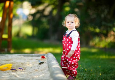 Dışarıda parkta kumlarda oynayan tatlı bir kız çocuğu. Kırmızı sakız pantolonlu güzel bebek güneşli yaz gününde eğleniyor. Renkli kum oyuncakları olan çocuk. Sağlıklı aktif bebek dışarıda oyunlar oynuyor.