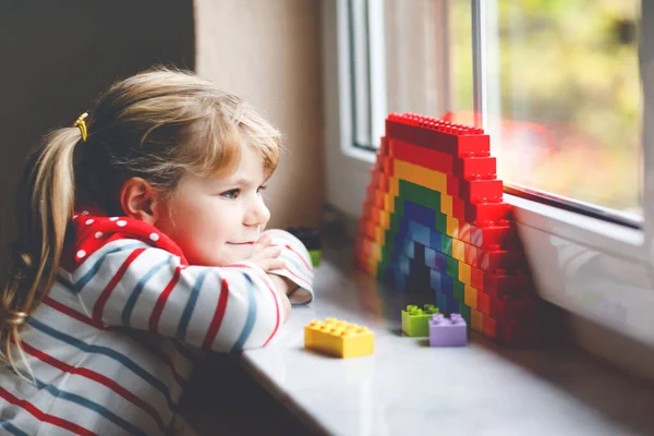 Schattig klein peuter meisje door het raam te creëren regenboog met kleurrijke plastic blokken tijdens pandemische coronavirus quarantaine. Kinderen maakten en schilderen regenbogen over de hele wereld als teken. — Stockfoto