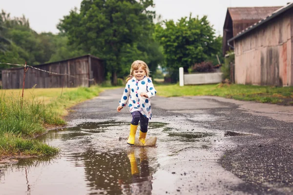 Kleines Mädchen in gelben Regenstiefeln, läuft und geht bei Schneeregen an einem regnerischen, bewölkten Tag. Nettes glückliches Kind in bunten Kleidern, das in eine Pfütze springt, mit Wasser planscht, Outdoor-Aktivität — Stockfoto