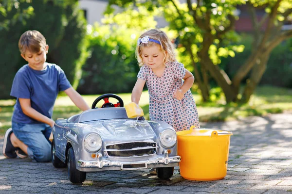 Zwei glückliche Kinder waschen im Sommergarten im Freien ein großes altes Spielzeugauto. Bruder Junge und kleine Schwester Kleinkind Mädchen reinigen Auto mit Seife und Wasser, haben Spaß beim Planschen und Spielen mit Schwamm. — Stockfoto