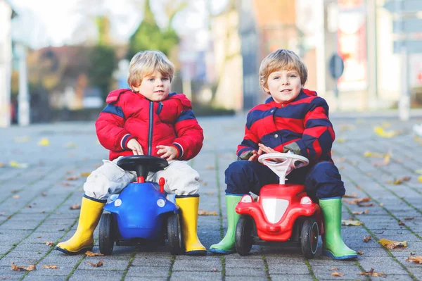 Zwei kleine Jungen in bunten Klamotten und Regenstiefeln fahren Spielzeugautos. Beste Freunde beim Wettstreit, im Freien. Aktive Freizeit für Kinder am Herbsttag. — Stockfoto