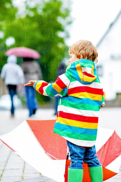 Little blond kid boy walking with big umbrella outdoors on rainy day. Child having fun with rain drops and wearing colorful waterproof clothes and boots — Stock Photo, Image