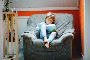 Pijamalı, şirin sarışın, anaokulu çocuğu. Ev odasında çocuklar için kitap okuyor. Heyecanlı çocuk yüksek sesle okuyor, eski büyük koltukta oturuyor. Okul çocuğu, aile, eğitim