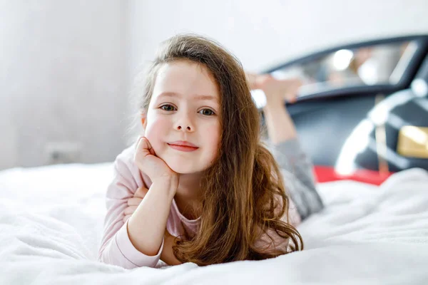 Imádnivaló, boldog kislány hosszú hajjal, miután a fehér szállodai ágyában aludt színes hálóingben. Iskolásgyerek családi vakációkon. Vicces boldog gyermek játszik és mosolyog — Stock Fotó