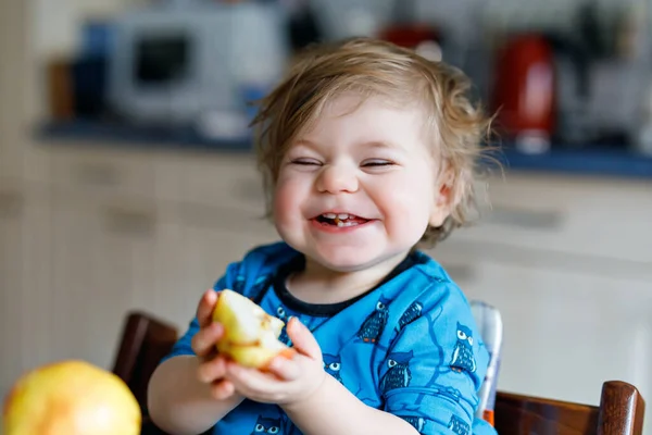 Menina adorável bonito bebê comendo pêra fresca. Criança feliz com fome de um ano segurando frutas. Menina na cozinha doméstica, tendo refeição saudável lanche. Criança loira sorrindo em casa ou no berçário — Fotografia de Stock