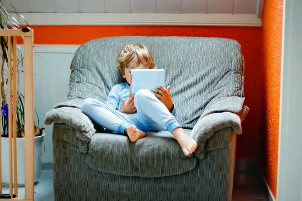 Cute blond little preschool kid boy in pajamas reading book for children in domestic room. Excited child reading loud, sitting on old big armchair. Schoolkid, family, education — Stock Photo, Image