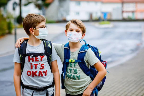 Dva chlapci s lékařskými maskami a brašnami. Školáci jsou na cestě do školy. Děti nosí ochrannou masku v důsledku kovidové pandemie viru korony. Nejlepší přátelé, bratři zpátky do školy po uzavření — Stock fotografie