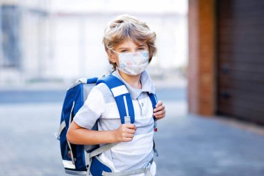 Happy little kid boy with medical mask and satchel walk to school. Schoolkid on way to school after lockdown. Healthy child outdoors. Back to school. Closed school due to corona virus covid pandemic