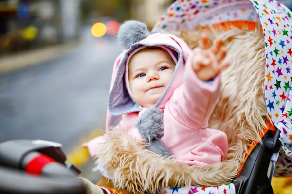 Cute little beautiful baby girl sitting in the pram or stroller on autumn day. Happy smiling child in warm clothes, fashion stylish pink baby coat with bunny ears. Baby going on a walk with parents. — Stock Photo, Image