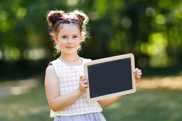 Happy little kid girl holding empty chalk desk in hands. Schoolkid on first day of elementary class. Healthy adorable child outdoors, in green park. Empty desk for copyspace text — Stockfoto