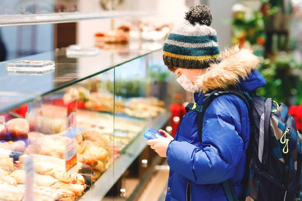 Een jongen met een medisch masker koopt brood en gebak voor de schoollunch in de bakkerij. Kind met rugzak en winterkleding. Schoolkind tijdens vergrendeling en quarantaine tijdens corona pandemische ziekte — Stockfoto