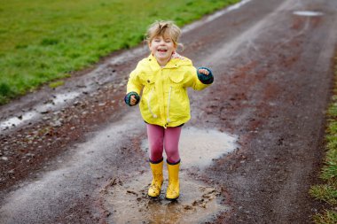 Little toddler girl wearing yellow rain gum boots, running and walking during sleet. Happy child in colorful clothes jumping into puddle, splashing with water, outdoor activity. Happiness, childhood