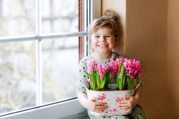 小さな幼児の女の子がピンクのヒヤシンスの花を咲かせて窓辺に座っています。幸せな子供室内で。母の日、バレンタインデー、誕生日、春のコンセプト. — ストック写真