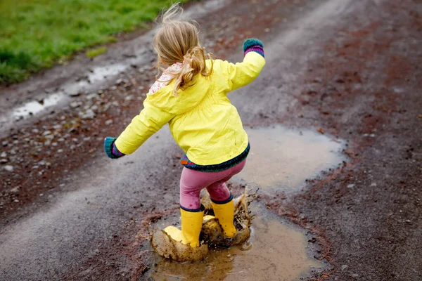 Little toddler girl wearing yellow rain gum boots, running and walking during sleet. Happy child in colorful clothes jumping into puddle, splashing with water, outdoor activity. Happiness, childhood — Stock Photo, Image