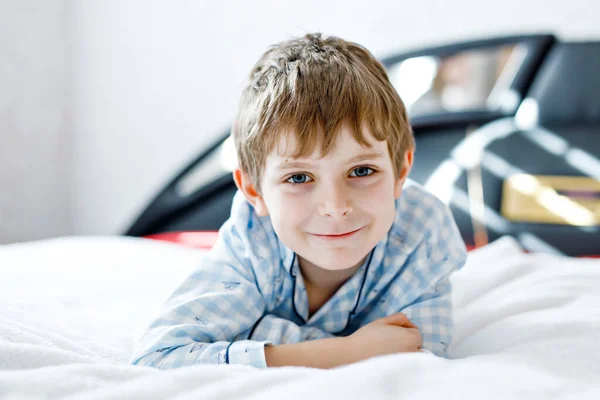 Roztomilý, šťastný chlapeček poté, co spal ve své bílé posteli v barevných nočních košilích. Školní dítě slaví pyžamovou párty a dívá se do kamery. Legrační šťastný dítě hrát a usmívat — Stock fotografie