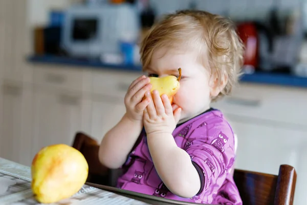 かわいい愛らしい幼児の女の子は新鮮な梨を食べています。1年間の果物を保持の空腹幸せな赤ちゃん。家庭用キッチンの女の子、健康的な食事の軽食を持っている。笑顔ブロンド幼児-ホームまたは保育園 — ストック写真